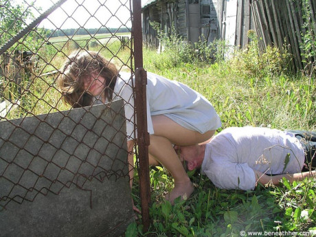 Caucasian lady GF broad straddles a boy BF man face outside of a doghouse in a yard - #173039