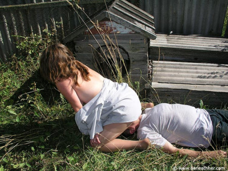 Caucasian lady GF broad straddles a boy BF man face outside of a doghouse in a yard - #173048