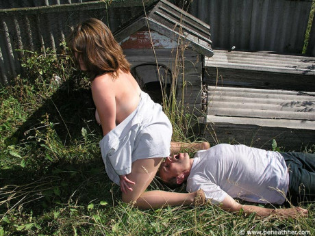 Caucasian lady GF broad straddles a boy BF man face outside of a doghouse in a yard - #173049